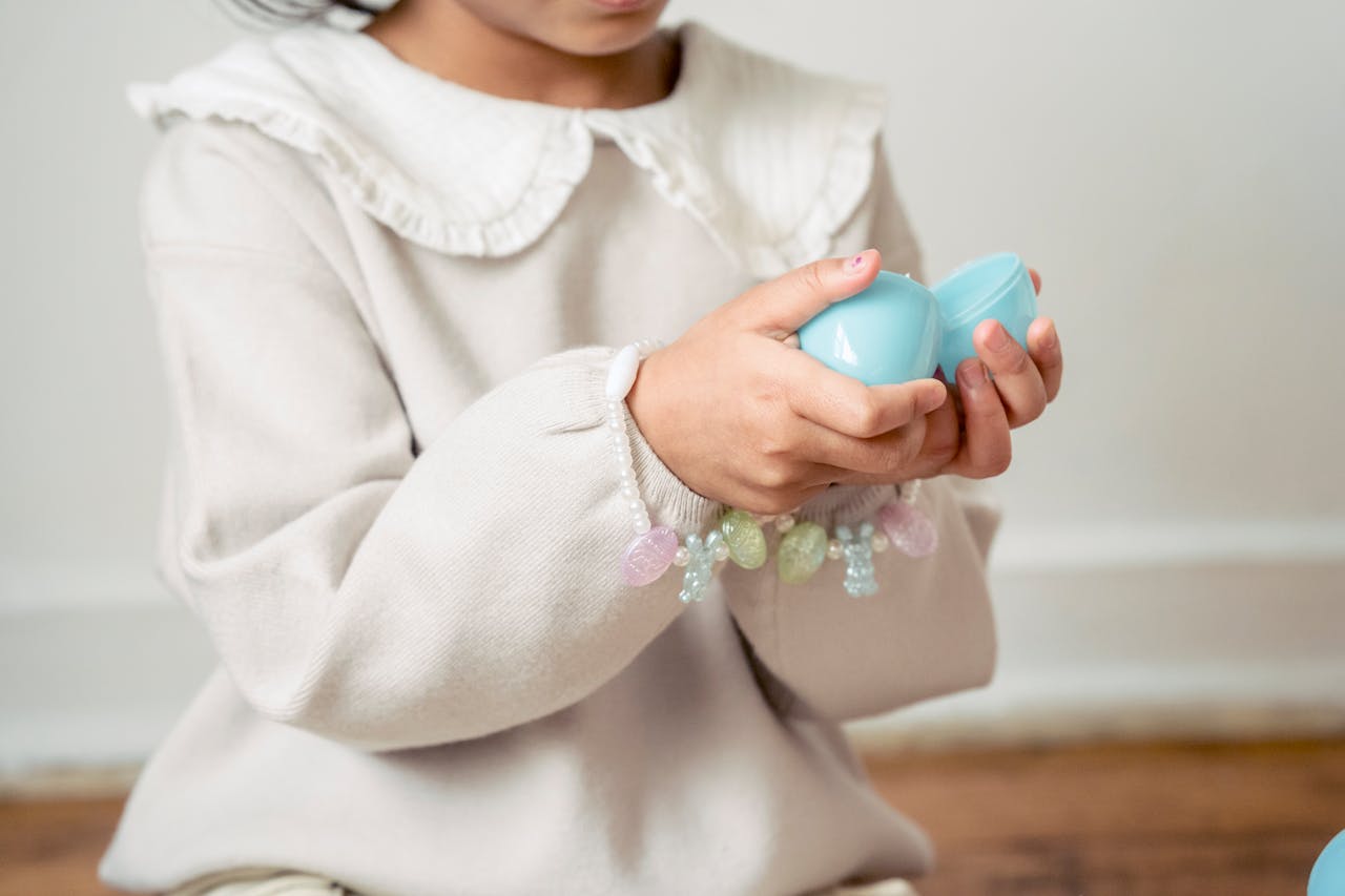 Crop unrecognizable girl in white dress opening surprise toy egg in light room