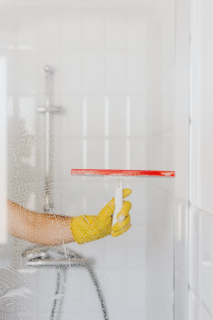 A person wearing a yellow glove cleans a shower glass with a squeegee, emphasizing hygiene and cleanliness.